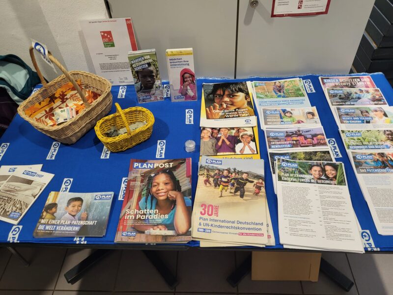 Auf einem mit einem blauen PLAN International-Tuch bedeckten Tisch liegen Broschüren, Zeitschriften und Flyer über Kinderpatenschaften und Kinderrechte aus. Zwei gelbe Körbe, ein QR-Code, Stifte und Werbematerialien sind ordentlich auf dem Tisch angeordnet.