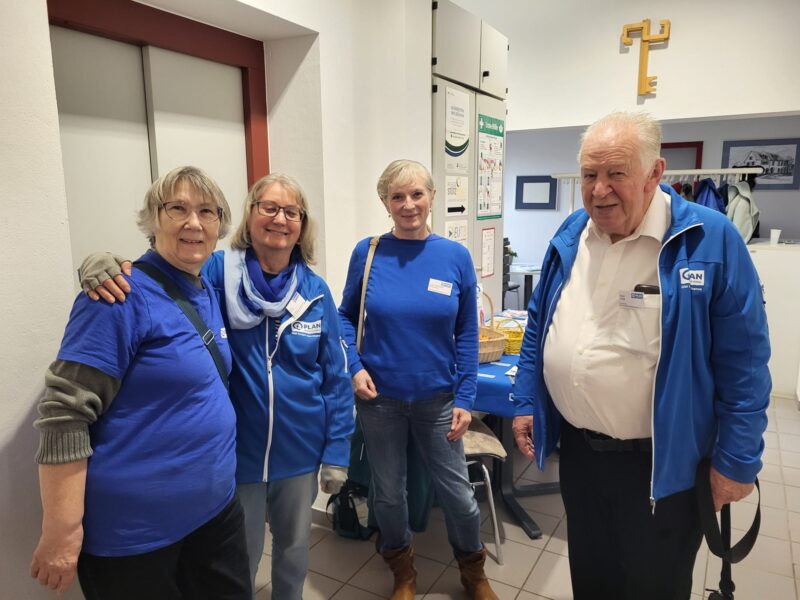 Vier ältere Erwachsene, drei Frauen und ein Mann, stehen in einem Haus und lächeln in die Kamera. Sie tragen blaue Kleidung mit Jacken und Namensschildern. Hinter ihnen befinden sich weiße Wände, eine Tür, Informationsplakate, ein Kleiderständer und eine große gelbe Wanddekoration in Form eines Schlüssels.