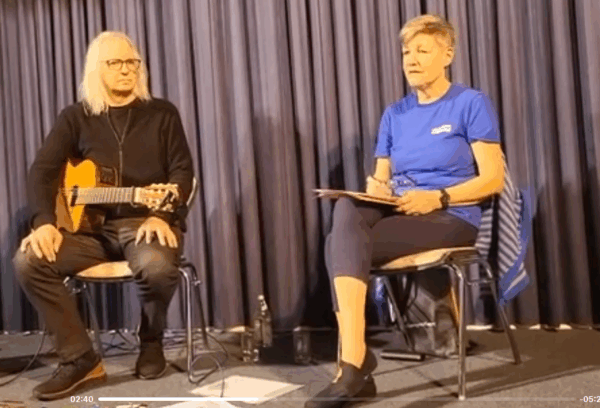 Zwei Personen sitzen auf Stühlen auf einer Bühne vor einem geschlossenen blauen Vorhang. Die Person auf der linken Seite hat langes weißes Haar, eine Brille, schwarze Kleidung und hält eine Akustikgitarre in der Hand. Die Person auf der rechten Seite trägt ein blaues Hemd, hält Papiere in der Hand und hat kurzes blondes Haar.