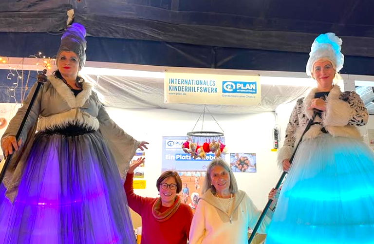 Zwei Frauen auf Stelzen tragen große, beleuchtete, elegante Kostüme - eine in Grau und die andere in Hellblau - und stehen neben zwei lächelnden Frauen in Mänteln. Sie posieren vor einem Stand mit einem Schild "PLAN Internationales Kinderhilfswerk".