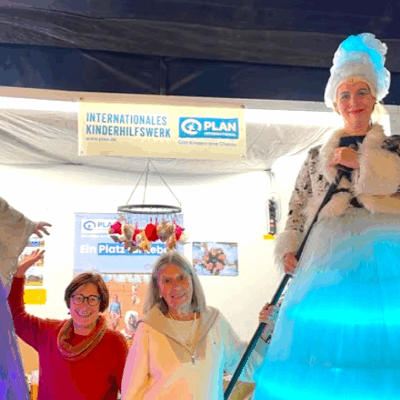 Vier Frauen posieren in einem Innenraum an einem Stand des Internationalen Kinderhilfswerks. Zwei Frauen tragen rote und weiße Pullover, lächeln und winken. Zwei andere stehen auf Stelzen und tragen aufwändige Kostüme, darunter ein langes blau-weißes Kleid und eine gepuderte Perücke.
