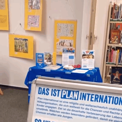 Auf einem mit einem blauen Plan International-Tuch bedeckten Tisch liegen Broschüren und Faltblätter aus. Dahinter hängen Kunstwerke von Kindern an einer Wand. Rechts daneben stehen Bücher in Regalen. Im Vordergrund wirbt ein Schild in deutscher Sprache für den Einsatz von Plan International für die Rechte der Kinder.