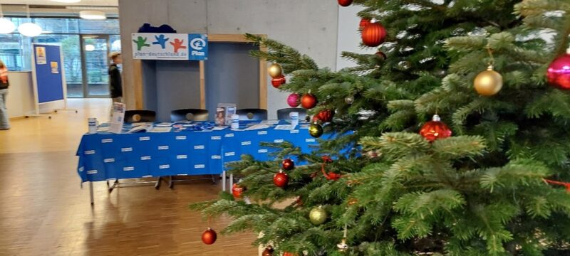 Ein geschmückter Weihnachtsbaum mit roten und goldenen Ornamenten steht in einem Innenraum neben einem blau gedeckten Tisch mit Broschüren und Flyern für eine Veranstaltung. Hinter dem Tisch sind bunte Handabdruckschilder und Informationsplakate zu sehen. Der Raum ist mit Holzböden und heller Beleuchtung ausgestattet.