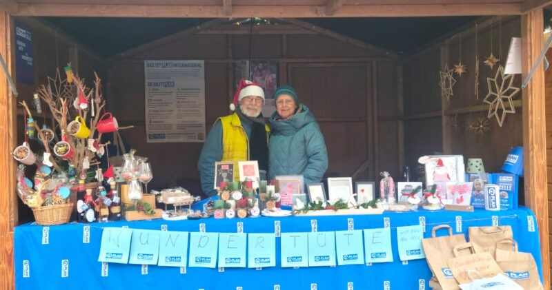 Zwei in Winterkleidung gekleidete Personen stehen in einem weihnachtlich geschmückten Holzstand. Der Tisch ist mit blauen Tüchern und handgefertigtem Kunsthandwerk, Geschenken und Backwaren bedeckt. Ein WUNDERTÜTE-Schild hängt über der Front. Ornamente und Dekorationen sind ausgestellt.