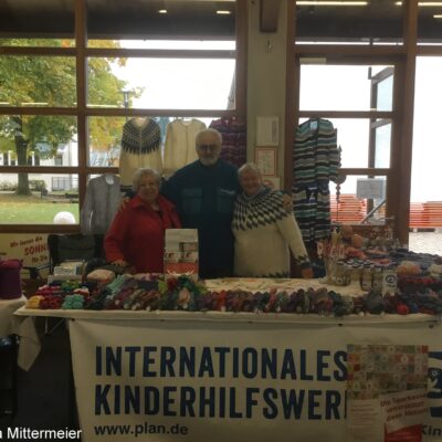 Drei ältere Erwachsene stehen lächelnd hinter einem Handwerksstand mit Garnprodukten und bunten gestrickten Socken. An dem Stand hängt ein Banner mit der Aufschrift "INTERNATIONALES KINDERHILFSWERK". Hinter ihnen hängen Pullover; durch große Fenster sind Bäume im Freien zu sehen.