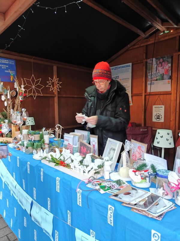 Eine Person mit roter Strickmütze und schwarzer Jacke arrangiert handgefertigtes Kunsthandwerk und Geschenke auf einem blauen Tischtuch an einem Marktstand im Freien. Der Stand bietet Kerzen, Ornamente, Fotos und kleine Dekorationen vor Holzwänden und festlichem Dekor im Hintergrund.