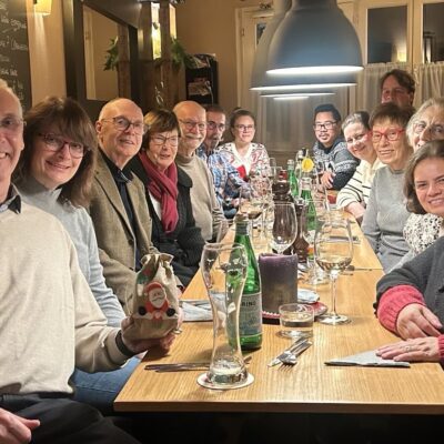 Eine Gruppe von fünfzehn Personen, Männer und Frauen unterschiedlichen Alters, sitzt an einem langen Holztisch in einem Restaurant und lächelt in die Kamera. Weingläser, Wasserflaschen und Besteck stehen auf dem Tisch. Die Umgebung wirkt warm und festlich.
