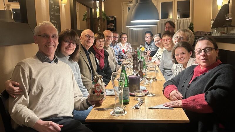 Eine Gruppe von fünfzehn Personen, Männer und Frauen unterschiedlichen Alters, sitzt an einem langen Holztisch in einem Restaurant und lächelt in die Kamera. Weingläser, Wasserflaschen und Besteck stehen auf dem Tisch. Die Umgebung wirkt warm und festlich.
