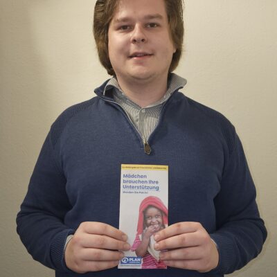 Ein Mann mit mittellangem braunem Haar, der einen blauen Pullover mit Reißverschluss über einem gestreiften Hemd trägt, steht in einem Innenraum vor einer beigen Wand. In der Hand hält er ein Flugblatt mit dem Foto eines lächelnden Mädchens und dem deutschen Text "Mädchen brauchen Ihre Unterstützung".