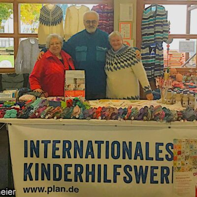 Drei lächelnde Menschen stehen hinter einem Tisch, der mit bunten Stricksachen und Garn bedeckt ist. Auf dem Tisch steht ein Schild mit der Aufschrift "Internationales Kinderhilfswerk" (International Childrens Aid Organization). Im Hintergrund sind ein Fenster und Bäume zu sehen.