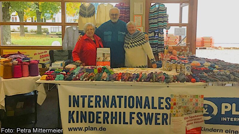 Drei lächelnde Menschen stehen hinter einem Tisch, der mit bunten Stricksachen und Garn bedeckt ist. Auf dem Tisch steht ein Schild mit der Aufschrift "Internationales Kinderhilfswerk" (International Childrens Aid Organization). Im Hintergrund sind ein Fenster und Bäume zu sehen.