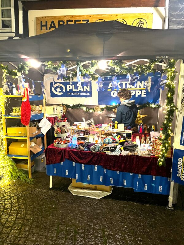 Ein nächtlicher Marktstand, geschmückt mit Lichterketten und festlichen Girlanden. Eine Person steht hinter einem Tisch, auf dem verschiedene bunte Artikel zum Verkauf angeboten werden. Im Hintergrund sind Banner von PLAN International und AKTIONS GRUPPE zu sehen. Der Boden ist mit Kopfsteinpflaster bedeckt.