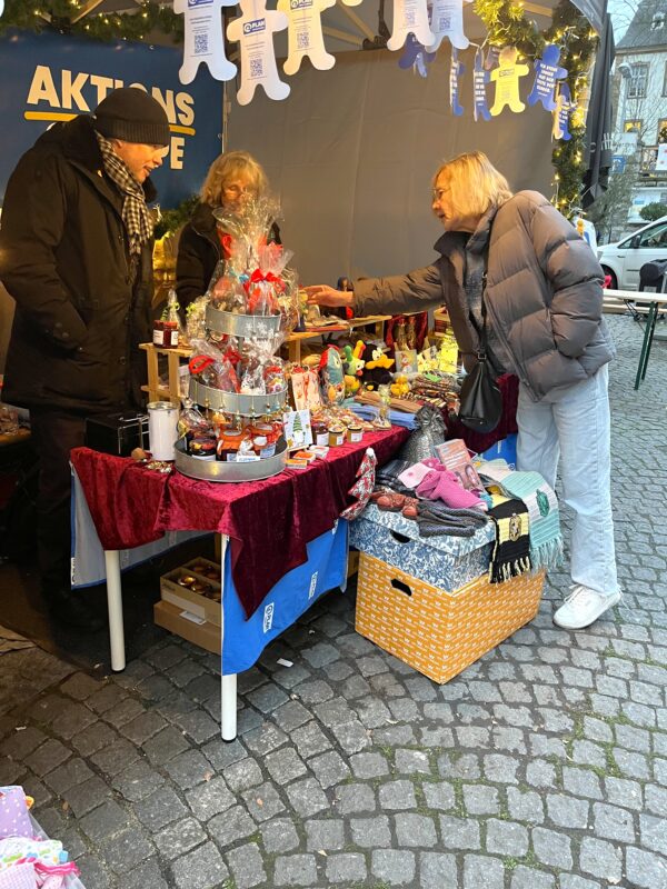Zwei Frauen und ein Mann stehen an einem Marktstand im Freien mit einem Tisch, auf dem Weihnachtsdekorationen, kleine Geschenke und gestrickte Artikel ausgestellt sind. Die Szene ist festlich mit Lichtern und Bannern oben und Kopfsteinpflaster unten. Eine Frau greift nach dem Verkäufer.