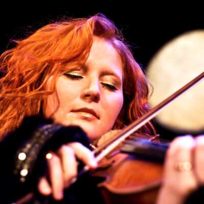 Eine Frau mit lockigem rotem Haar spielt mit geschlossenen Augen Geige und ist in die Musik vertieft. Sie trägt ein schwarzes Oberteil und ist warm beleuchtet, mit einem verschwommenen runden Licht im Hintergrund vor einer dunklen Kulisse.