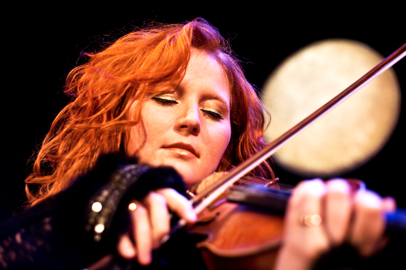 Eine Frau mit lockigem rotem Haar spielt mit geschlossenen Augen Geige und ist in die Musik vertieft. Sie trägt ein schwarzes Oberteil und ist warm beleuchtet, mit einem verschwommenen runden Licht im Hintergrund vor einer dunklen Kulisse.