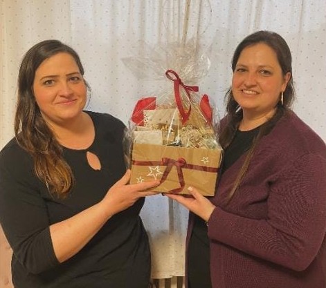 Zwei Frauen mit langen dunklen Haaren stehen in einem Haus nebeneinander und lächeln in die Kamera. Sie halten einen festlichen, in Zellophan eingewickelten und mit rotem Band und Sternenmustern verzierten Geschenkkorb in der Hand. Im Hintergrund ist ein weißer Vorhang mit kleinen Punkten zu sehen.