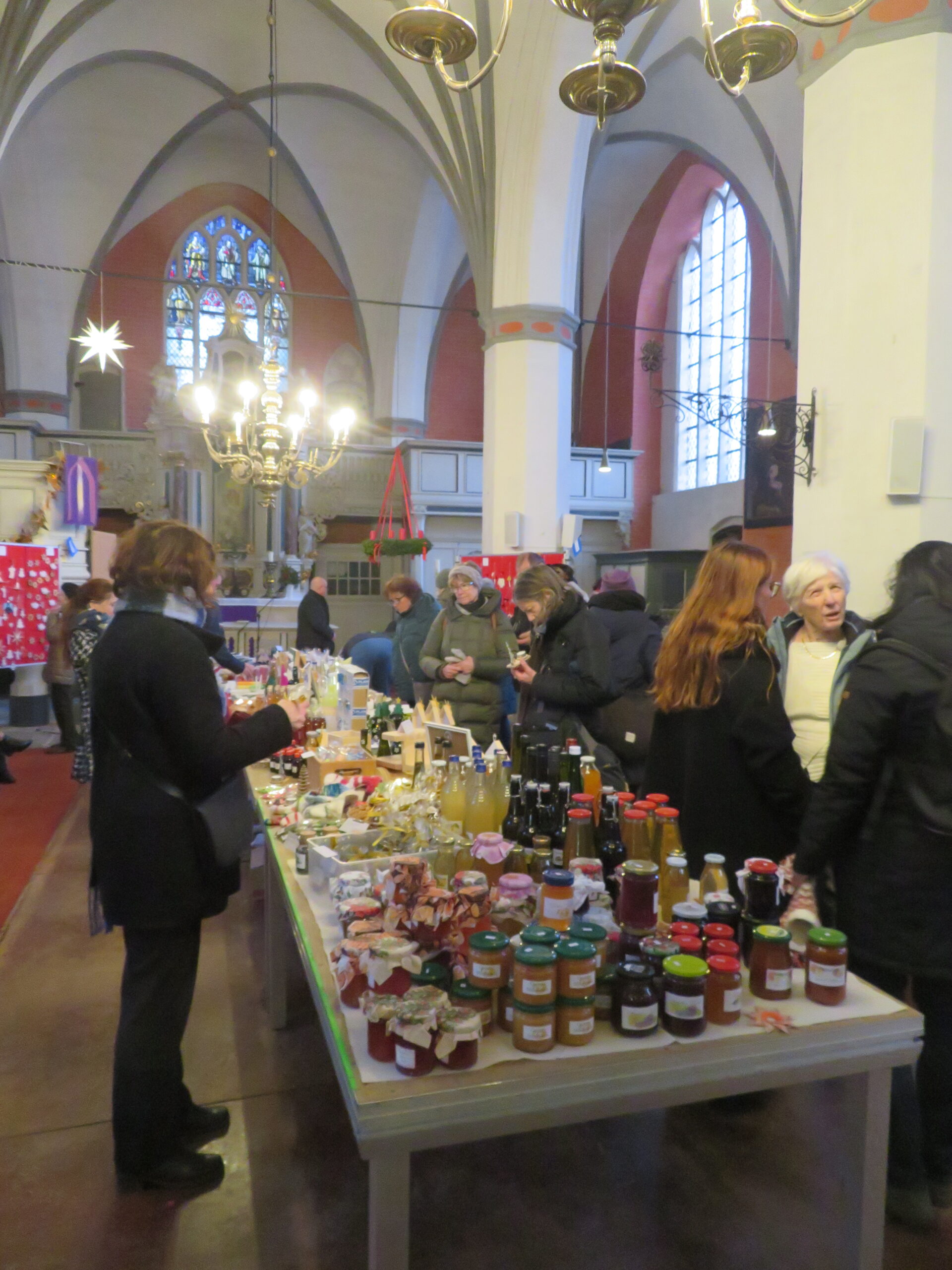 Auf einem Markt in einer Kirche mit hohen Gewölbedecken, Kronleuchtern und Buntglasfenstern stöbern die Menschen auf Tischen, die mit Einmachgläsern, Flaschen und handgefertigten Waren gefüllt sind. Die Atmosphäre ist lebhaft, und Gruppen unterhalten sich in der Nähe der Tische.