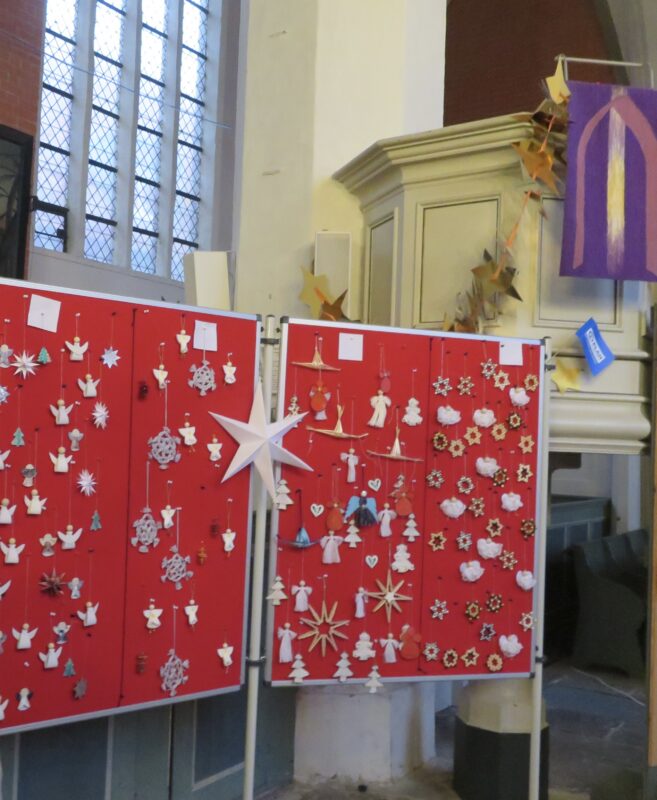 In einer Kirche hängt auf roten Tafeln handgefertigter Weihnachtsschmuck. Die Ornamente haben die Form von Sternen, Engeln, Schneeflocken und Bäumen. Im Hintergrund sind Buntglasfenster und eine mit Papiersternen und einem lila Tuch geschmückte Kanzel zu sehen.