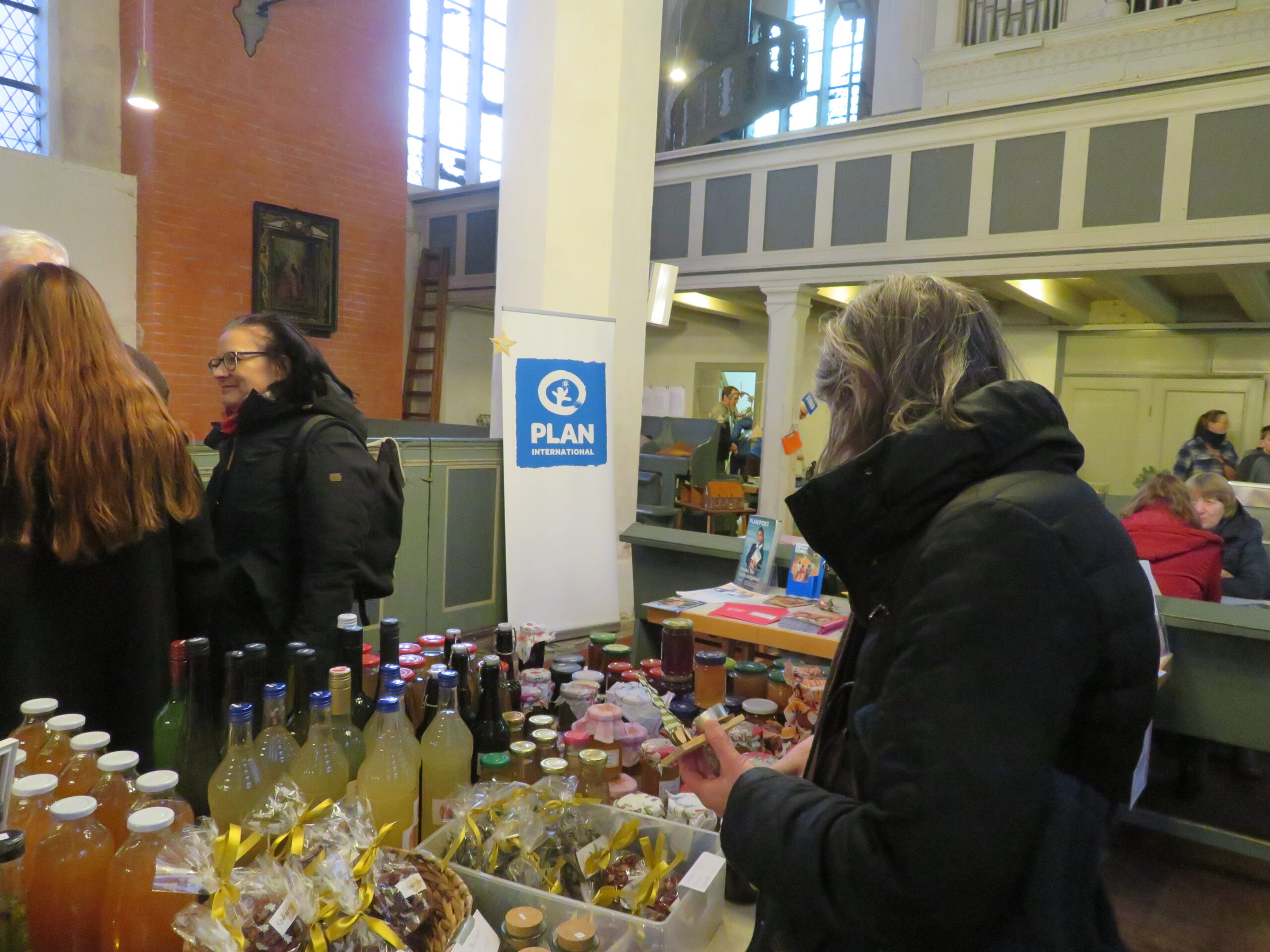 Eine Frau in einem schwarzen Mantel betrachtet Gläser und Flaschen mit selbstgemachten Waren auf einem Tisch in einem Haus. Andere Leute stöbern in der Nähe. Hinter ihnen befindet sich ein blaues PLAN International-Schild, Bänke, Stände und hohe Fenster in einer hellen, geräumigen Halle.