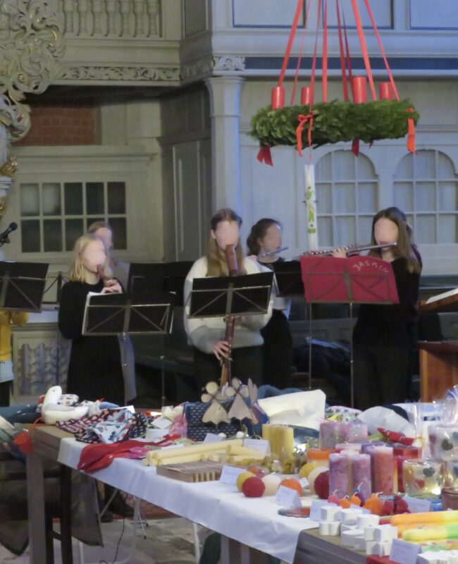 Vier Musiker mit verschwommenen Gesichtern spielen drinnen Blasinstrumente in der Nähe eines Tisches voller farbenfroher handgefertigter Gegenstände, Kerzen und Weihnachtsartikel. Über ihnen hängt ein großer Adventskranz mit vier roten Kerzen, was auf eine festliche, weihnachtliche Atmosphäre hindeutet.