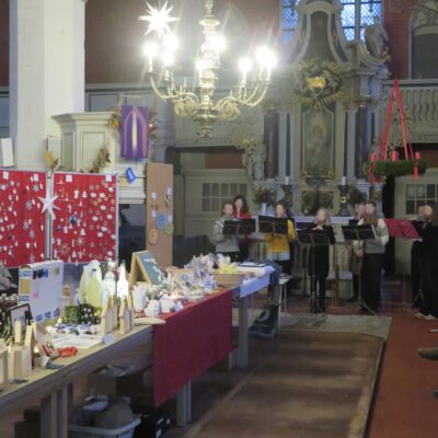 Ein festlicher Markt in einer Halle mit Tischen mit handgefertigtem Kunsthandwerk und Geschenken. Die Leute stöbern in Auslagen mit Schmuck, Kerzen und Dekorationen. Im Hintergrund spielen Musiker Blasinstrumente in der Nähe eines verzierten Altars. Der Raum wird von einem großen Kronleuchter über dem Kopf beleuchtet.