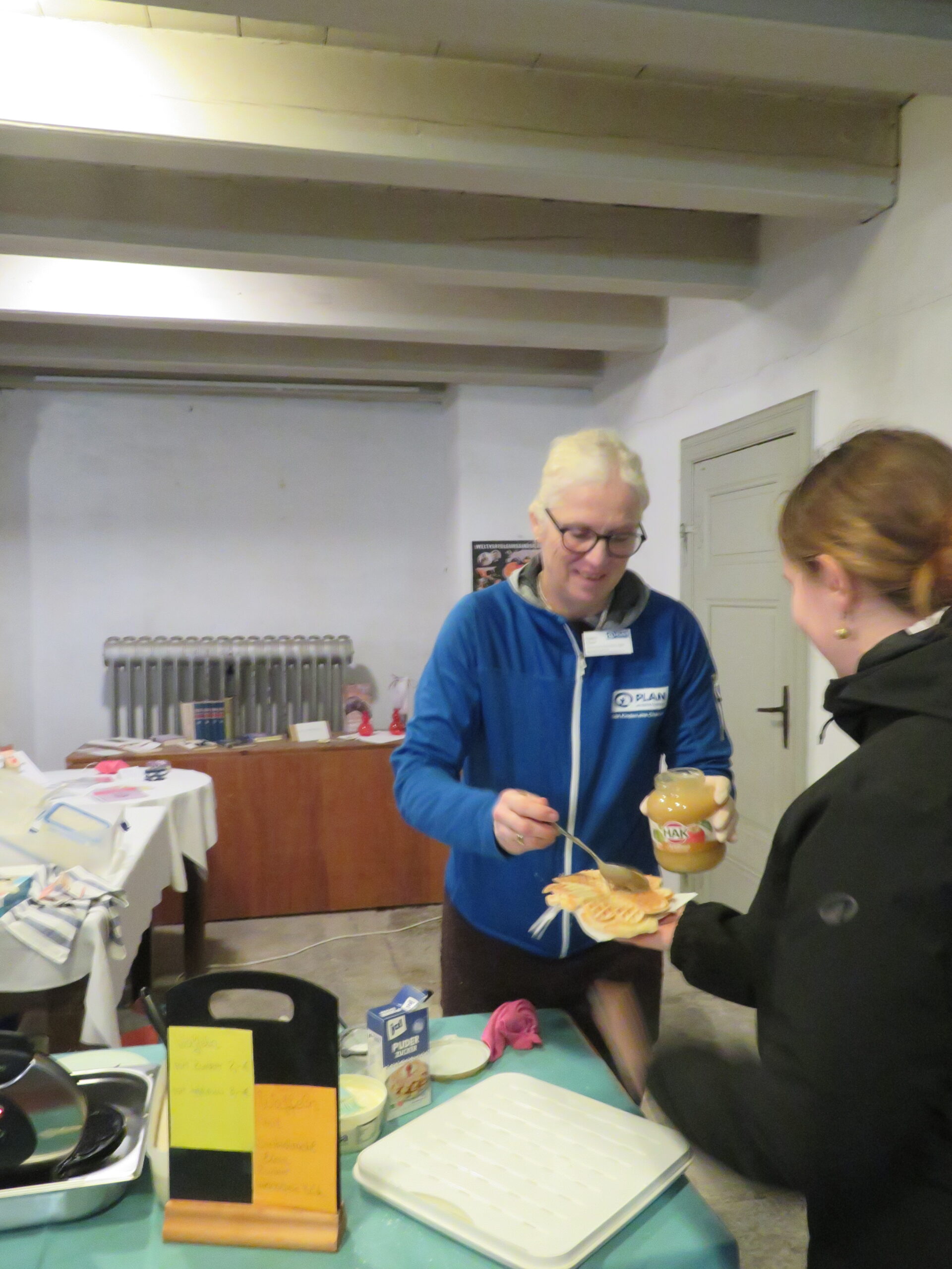 Eine lächelnde Person mit Brille und weißem Haar, die eine blaue Jacke und ein Namensschild trägt, verteilt Erdnussbutter auf einer Waffel für eine andere Person an einem Tisch. Auf dem Tisch befinden sich ein Zettel, Utensilien und Gläser. Im Hintergrund sind ein Heizkörper und weitere Tische mit verschiedenen Gegenständen zu sehen.