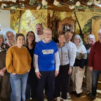 Eine Gruppe von zwölf Erwachsenen, Männern und Frauen, steht in einem Innenraum dicht beieinander und lächelt in die Kamera. Ein Mann im Vordergrund trägt ein blaues Hemd mit der Aufschrift "AKTIONS GRUPPE". Im Hintergrund sieht man rustikale Holzbalken, eine Wandmalerei aus Laub und eine warme, gemütliche Beleuchtung.