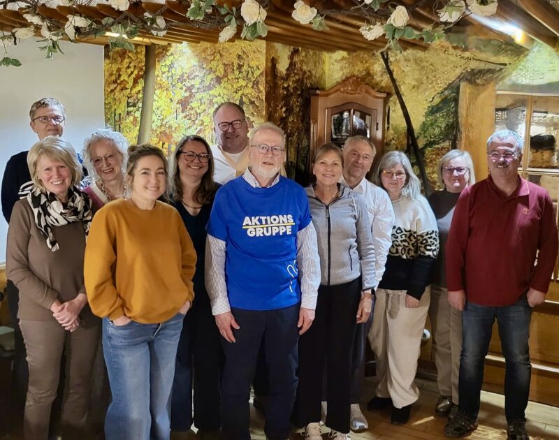 Eine Gruppe von zwölf Erwachsenen, Männern und Frauen, steht in einem Innenraum dicht beieinander und lächelt in die Kamera. Ein Mann im Vordergrund trägt ein blaues Hemd mit der Aufschrift "AKTIONS GRUPPE". Im Hintergrund sieht man rustikale Holzbalken, eine Wandmalerei aus Laub und eine warme, gemütliche Beleuchtung.