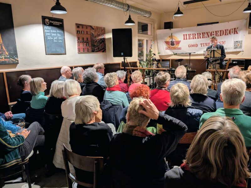 Eine Gruppe älterer Menschen sitzt vor einer Bühne in einem gemütlichen Café. Ein Mann steht an einem Mikrofon unter einem Transparent mit der Aufschrift "Herrengedeck Good Time Music". Auf der Bühne stehen Musikinstrumente und Lautsprecher; warmes Licht schafft eine einladende, aufmerksame Atmosphäre.