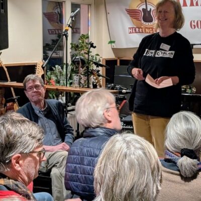 Eine Frau steht lächelnd da und spricht zu einem sitzenden Publikum in einem gemütlichen Innenbereich, möglicherweise einem Café oder einem kleinen Veranstaltungsort. Im Hintergrund sind Musikinstrumente und ein Banner mit einem Gitarrenlogo zu sehen, und das Publikum besteht hauptsächlich aus älteren Erwachsenen.