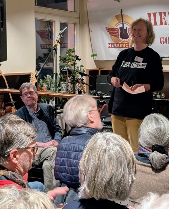 Eine Frau steht lächelnd da und spricht zu einem sitzenden Publikum in einem gemütlichen Innenbereich, möglicherweise einem Café oder einem kleinen Veranstaltungsort. Im Hintergrund sind Musikinstrumente und ein Banner mit einem Gitarrenlogo zu sehen, und das Publikum besteht hauptsächlich aus älteren Erwachsenen.