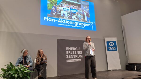 Eine Frau steht auf der Bühne, spricht in ein Mikrofon und hält Papiere in der Hand. Zwei Frauen sitzen links von ihr und hören zu. Hinter ihr ist ein Bildschirm mit einem Gruppenfoto und der Aufschrift "Plan-Aktionsgruppen" zu sehen. "ENERGIE ERLEBNIS ZENTRUM" und ein Transparent von Plan International sind zu sehen.