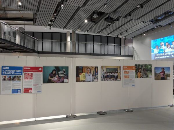 Ausstellungsraum mit einer Reihe von Tafeln, auf denen Poster und Fotos zum Thema Gleichstellung ausgestellt sind. Die Tafeln zeigen Menschen bei der Arbeit, beim Studium und Familienszenen. An der Wand ist ein Willkommensgruß in deutscher Sprache projiziert. Darüber sind ein moderner Balkon und eine Decke mit sichtbaren Balken zu sehen.