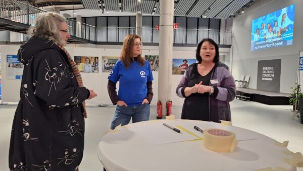 Drei Frauen stehen um einen runden Tisch mit Papier, Stiften und Klebeband in einem großen, modernen Raum. Eine Frau trägt einen schwarz gemusterten Mantel, eine andere ein blaues Hemd, und die dritte gestikuliert beim Sprechen. Im Hintergrund sind Ausstellungsdisplays und ein großer Bildschirm zu sehen.