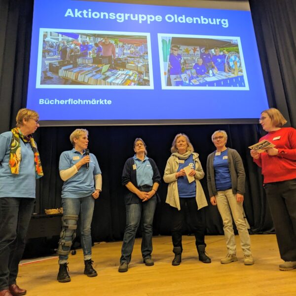 Fünf Frauen stehen auf der Bühne unter einer Leinwand mit der Aufschrift Aktionsgruppe Oldenburg, zwei Fotos und dem Wort Bücherflohmarkte. Eine Frau in Rot hält ein Mikrofon, während die anderen aufmerksam zuhören. Alle tragen Namensschilder und Freizeitkleidung.