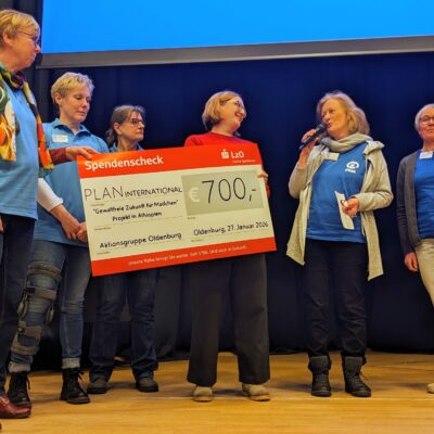 Sechs Frauen stehen auf einer Bühne. Vier von ihnen halten einen großen Scheck über 700 Euro, ausgestellt auf PLAN International. Eine Frau spricht in ein Mikrofon. Alle tragen Freizeitkleidung und Namensschilder und wirken glücklich. Hinter ihnen ist eine Projektionsfläche zu sehen.