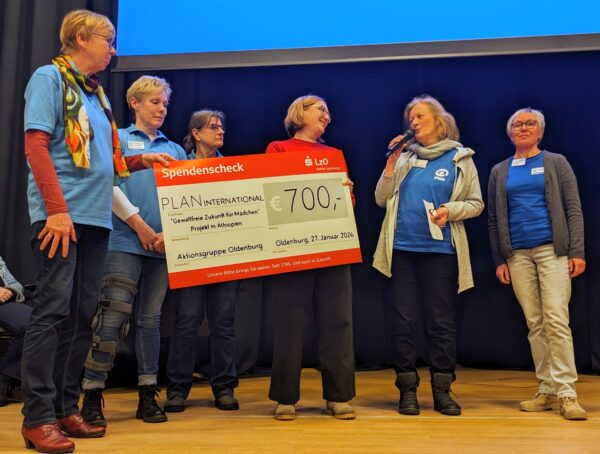 Sechs Frauen stehen auf einer Bühne. Vier von ihnen halten einen großen Scheck über 700 Euro, ausgestellt auf PLAN International. Eine Frau spricht in ein Mikrofon. Alle tragen Freizeitkleidung und Namensschilder und wirken glücklich. Hinter ihnen ist eine Projektionsfläche zu sehen.