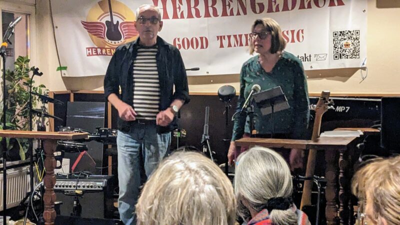 Zwei Personen stehen auf einer kleinen Bühne vor einem Publikum. Hinter ihnen ist ein Banner mit der Aufschrift "Herzengel..." und musikalischen Grafiken zu sehen. Der Mann trägt eine Brille, ein gestreiftes Hemd und eine Jeans. Die Frau trägt eine Brille und ein grünes Kleid. Mikrofone und Musikgeräte sind zu sehen.