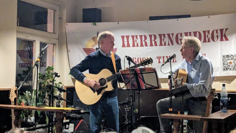 Zwei Männer spielen Akustikgitarren und singen in Mikrofone in einer kleinen Halle. Einer steht, einer sitzt. Hinter ihnen hängt ein Transparent mit der Aufschrift HERRENDECK. In dem gemütlichen, warm beleuchteten Raum sind Topfpflanzen, Notenständer und Audiogeräte zu sehen.