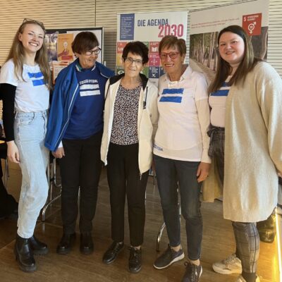 Fünf Frauen stehen lächelnd in einem Haus zusammen. Vier tragen blau-weiße Hemden, eine trägt eine gemusterte Bluse mit weißer Jacke. Hinter ihnen hängen Plakate über die Agenda 2030 und die UN-Ziele für nachhaltige Entwicklung. Die Atmosphäre ist locker und freundlich.