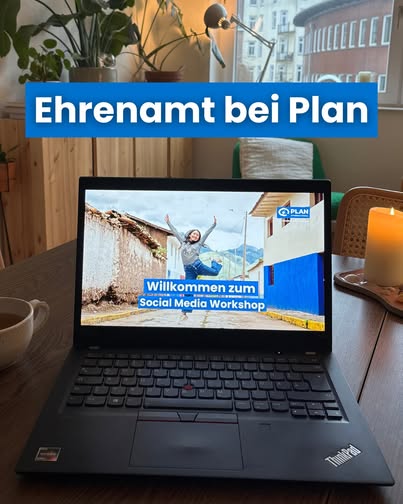 Ein Laptop auf einem Tisch zeigt das Bild einer lächelnden Person mit erhobenen Armen im Freien und dem Text Willkommen zum Social Media Workshop. Über dem Laptop steht in fetter Schrift "Ehrenamt bei Plan". In der Nähe stehen eine Kerze, eine Tasse und Pflanzen.