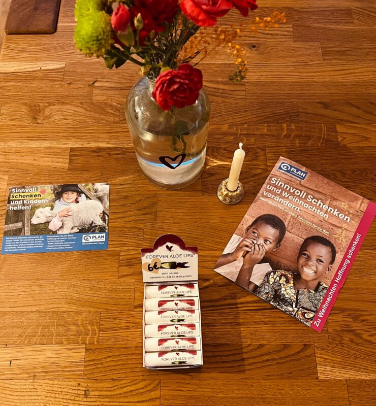 Ein Holztisch mit einer Vase mit roten Blumen, einer brennenden Kerze, einer Broschüre von Plan International mit zwei lächelnden Kindern, einer Postkarte und einer Pappschachtel mit sechs Forever Aloe Lips-Balsamen. Der Tisch ist warm beleuchtet, was eine gemütliche Atmosphäre schafft.