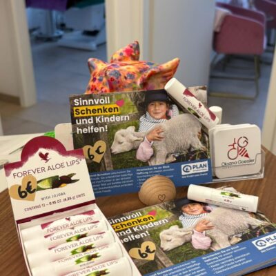 Ein Tisch mit einer Schachtel Forever Aloe Lippenbalsam, zwei Werbebroschüren mit einem Kind und einem Schaf, einem Holzherz, einem bunten Spielzeugstern und einem stehenden Schild mit einem roten Logo. Die Umgebung scheint eine medizinische oder Büroumgebung zu sein.