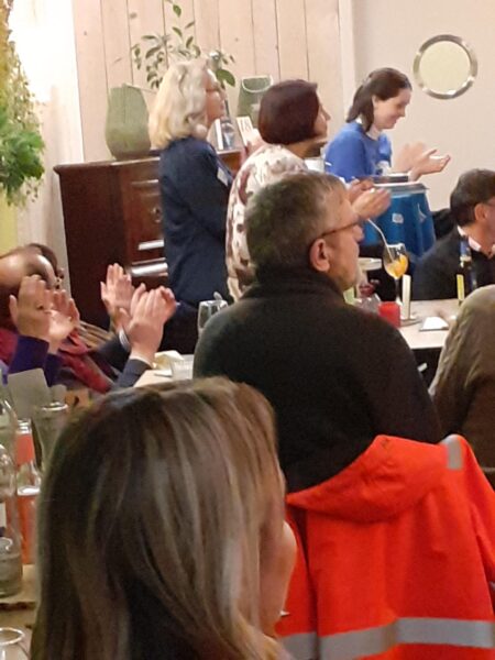 Eine Gruppe von Menschen ist in einem gemütlichen Innenraum versammelt, einige sitzen an Tischen mit Getränken und Gläsern, während andere stehen und applaudieren. Die Atmosphäre wirkt warm und gesellig, mit Pflanzen und Holzdekor im Hintergrund.