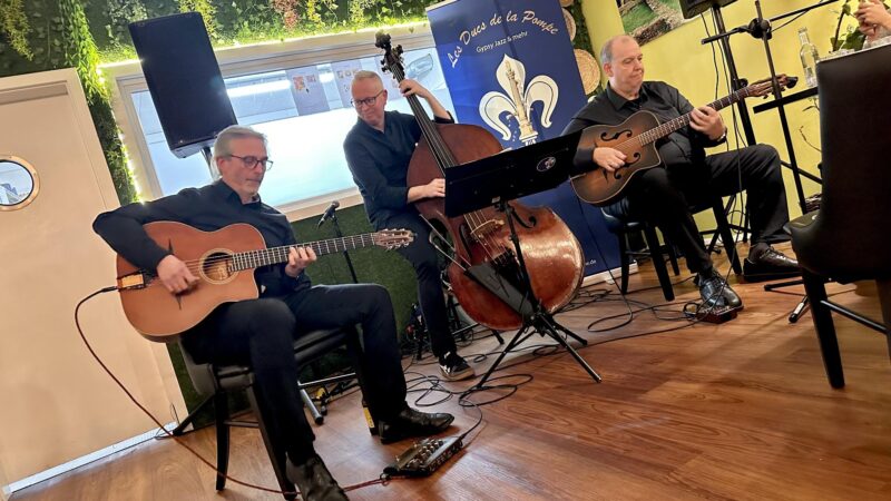 Drei schwarz gekleidete Musiker treten in einem Innenraum auf: zwei spielen Gitarren und einer einen Kontrabass. Sie sitzen neben einem blauen Banner mit einer Lilienblüte und "Duits de la Poete". Um sie herum sind auf einem Holzfußboden Notenständer und Mikrofone aufgebaut.