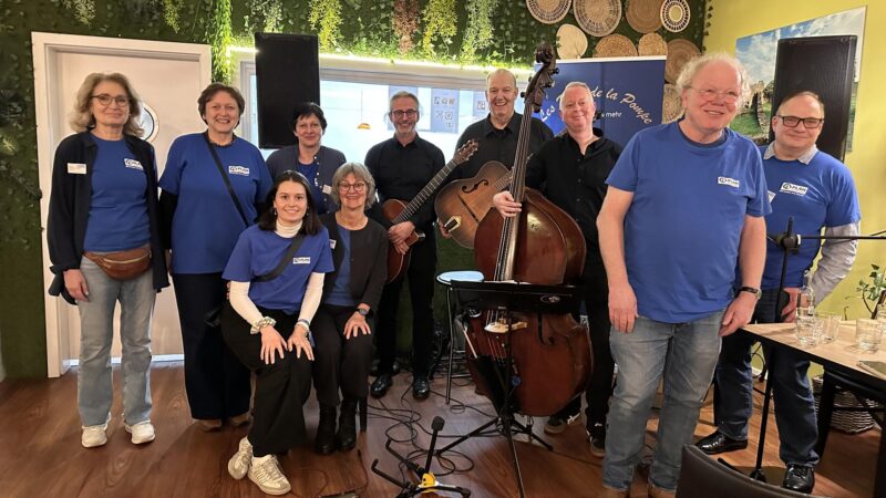 Eine Gruppe von elf Personen, von denen einige passende blaue Hemden tragen, posiert in einem Innenraum. In der Mitte stehen vier schwarz gekleidete Musiker - mit einem Doppelbass -. Die Umgebung ist hell, mit grünen Pflanzen und Bilderrahmen an der Wand und Gläsern auf einem Holztisch in der Nähe.