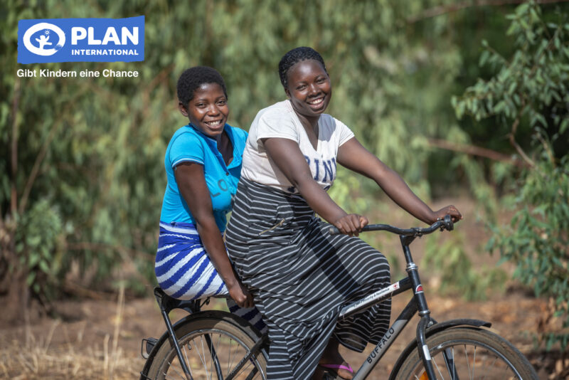 Zwei lächelnde Mädchen in gestreiften Röcken fahren zusammen auf einem Fahrrad im Freien, mit grünem Laub im Hintergrund. Das Bild zeigt das Logo von Plan International und den deutschen Satz Gibt Kindern eine Chance in der oberen linken Ecke.