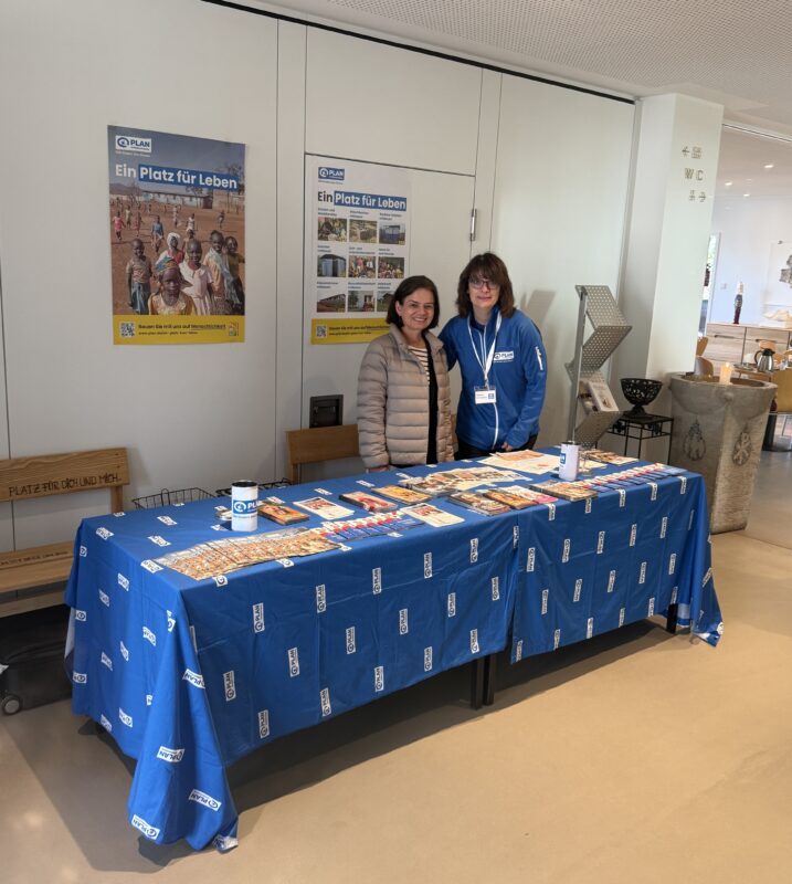Zwei Frauen stehen hinter einem mit einem blauen Tischtuch bedeckten Infotisch mit Broschüren, Flyern, Stiften und Tassen. Hinter ihnen befinden sich Plakate mit deutschen Texten und Fotos von Menschen. Die Umgebung scheint ein öffentlicher Raum oder eine Lobby zu sein.