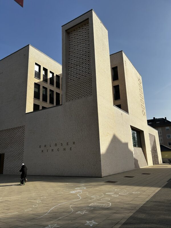 Ein modernes, helles Backsteingebäude mit der Aufschrift "Erlöser Kirche" steht unter einem klaren blauen Himmel. Das Sonnenlicht wirft Schatten auf die Fassade. Kreidezeichnungen von Sternen, Linien und Formen zieren den Boden, und eine Person fährt mit einem Motorroller vorbei.