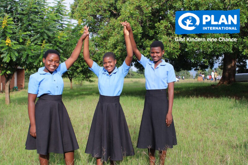 Drei lächelnde Mädchen in blauen Hemden und schwarzen Röcken stehen im Gras, zwei halten die Hände hoch. Sie befinden sich im Freien mit grünen Bäumen im Hintergrund. In der oberen rechten Ecke befindet sich das Logo von Plan International und der Text: Gibt Kindern eine Chance.